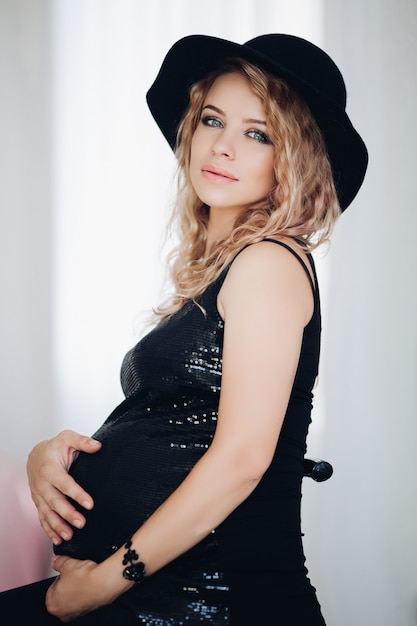 Stilvolle schwangere Frau im schwarzen Hut und in funkelnder Spitze, die ihren Bauch umfassen.