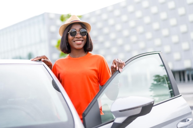 Stilvolle sommerliche schwarze Frau in orangefarbenem Hemd und Sonnenbrille öffnet lächelnd die Autotür