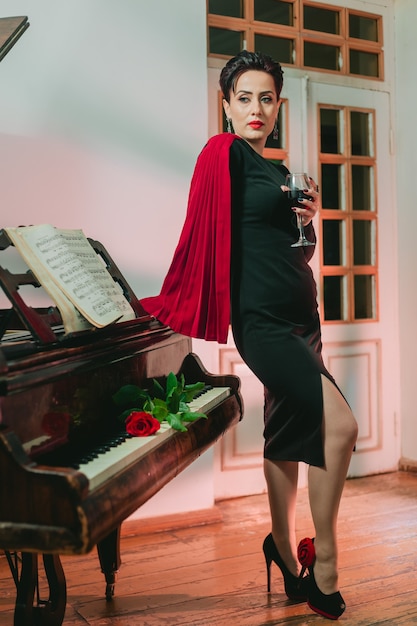 Stilvolle und elegante Frau mit kreativem rotem Mantel, der mit roter Rose auf dem Klavier steht und sich lehnt
