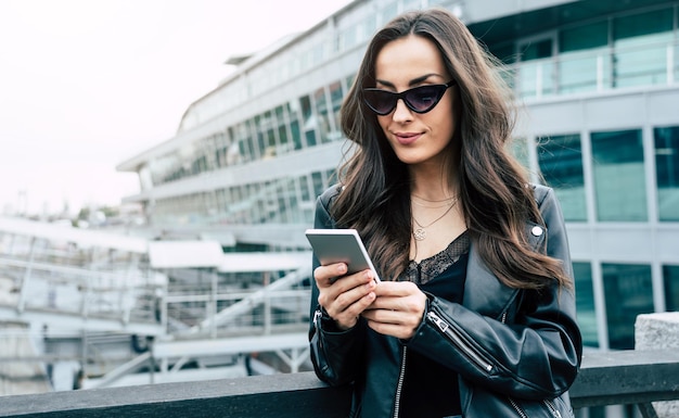 Stilvolle und trendige junge schöne Frau mit Sonnenbrille und im Hipster-Stil, während sie ein Telefon benutzt und Spaß im Freien in der Stadt hat