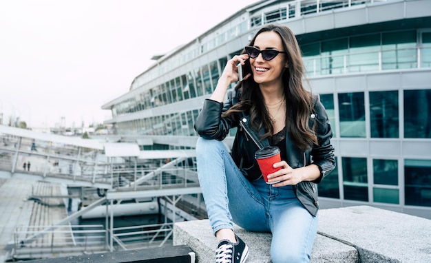 Stilvolle und trendige junge schöne Frau mit Sonnenbrille und im Hipster-Stil, während sie ein Telefon benutzt und Spaß im Freien in der Stadt hat