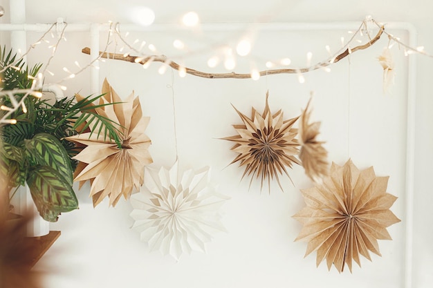 Foto stilvolle weihnachtssterne und -lichter aus papier, die am weißen wandhintergrund hängen. skandinavische festliche dekoration im boho-raum