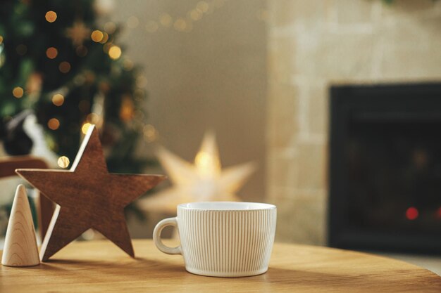 Stilvolle weiße Tasse mit hölzernem Sterndekor auf dem Tisch vor stilvollem, festlichem Weihnachtsbaum mit goldenen Lichtern und gemütlichem Kamin. Tassenattrappe für Weihnachtswerbeproduktvorlage. Platz für Text