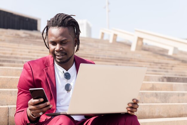 Stilvoller afrikanischer geschäftsmann, der im freien mit laptop und smartphone arbeitet