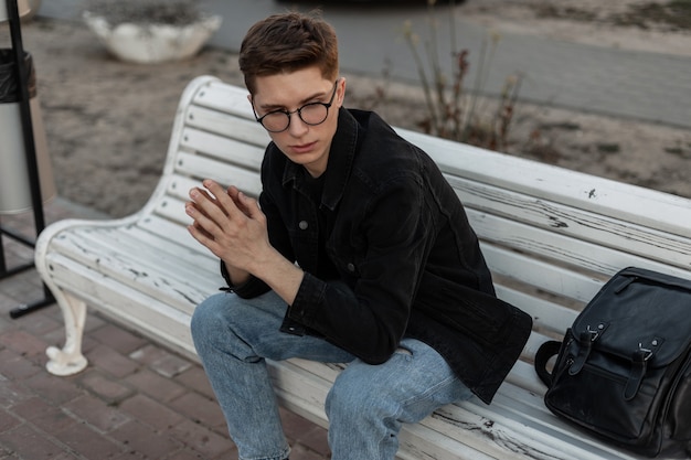 Stilvoller amerikanischer Mann in lässiger Denim-Kleidung mit schwarzem Lederrucksack sitzt auf einer Vintage-Holzbank in der Stadt. Attraktiver trendiger Kerl in modischer Jugendkleidung ruht auf der Straße.