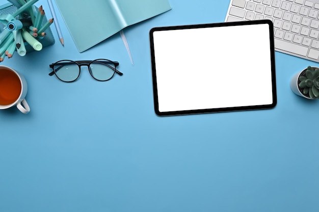 Stilvoller Arbeitsplatz der Draufsicht mit digitalem Tablet, Gläsern, Kaffeetasse und Notizbuch auf blauem Hintergrund.