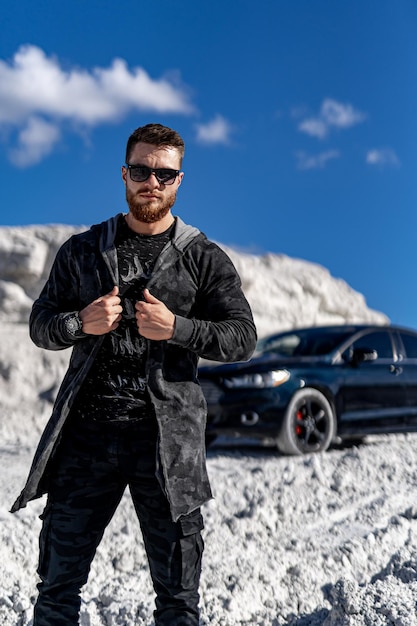 Stilvoller bärtiger Mann in trendiger Sonnenbrille und modischer Jacke steht in der Nähe eines schwarzen Autos. Modeporträt auf weißem Felsenhintergrund.