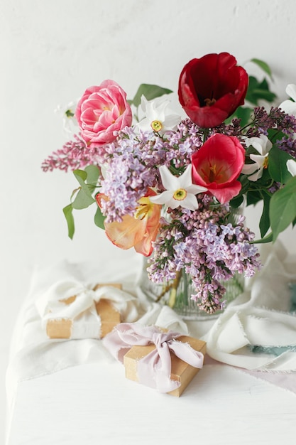 Stilvoller Blumenstrauß mit Geschenkbox auf rustikalem Holztisch. Fröhlicher Muttertag. Frühlingsgesteck aus lila Tulpen, Narzissen und Geschenk auf weichem Stoff in einem modernen, rustikalen Zimmer. Fröhlicher Frauentag