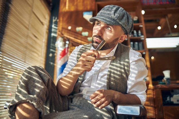 Stilvoller Friseur, der einen spitzen Hut trägt und nachdenklich aussieht, während er in einem Sessel sitzt und ein Rasiermesser hält