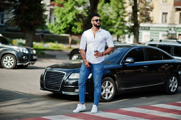 Stilvoller großer Mann im weißen Hemd, in den Jeans und in der Sonnenbrille, die an der Straße des Stadtbart-attraktiven Kerls mit Tasse Kaffee gegen schwarzes Geschäftsauto gestellt werden.