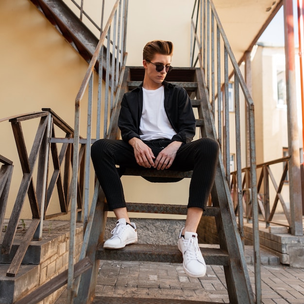 Stilvoller Hipster-Mann in Sonnenbrille in einem schwarzen Hemd in einem T-Shirt in eleganter gestreifter Hose in Turnschuhen sitzt auf einer eisernen Vintage-Treppe