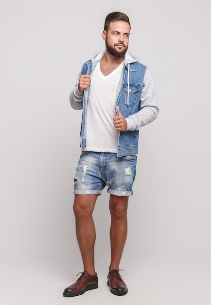 Stilvoller junger fröhlicher Kerl mit einem schönen Bart in einer Jeansjacke, einem weißen T-Shirt und Jeansshorts auf einem einfachen grauen Raum. Werbekonzept für Jeansjacke und Jeansshorts für Werbetafeln.