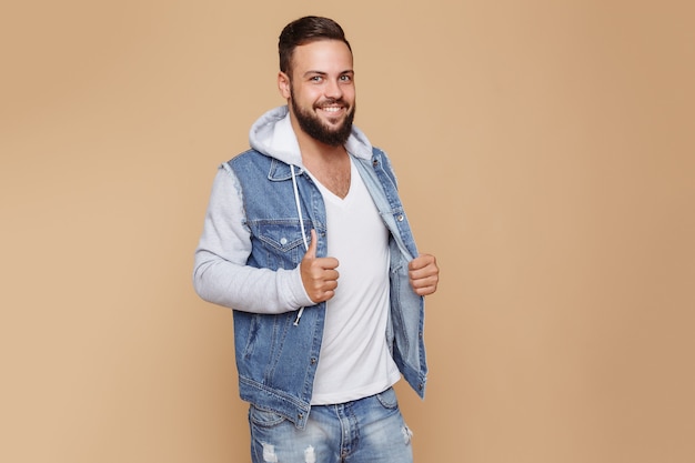 Stilvoller junger fröhlicher Kerl mit einem schönen Bart in einer Jeansjacke und weißem T-Shirt auf einem einfachen cremefarbenen Raum. Jeansjacke Werbekonzept für Plakatwand.