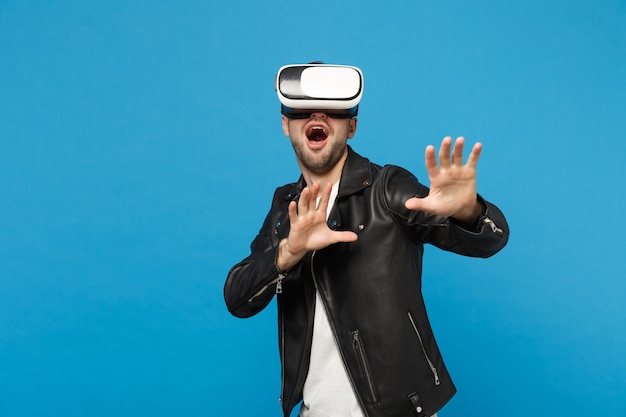 Stilvoller junger unrasierter Mann im weißen T-Shirt der schwarzen Jacke, der im Kopfhörer schaut, virtuelle VR-Realität einzeln auf blauem Wandhintergrundstudioporträt. Menschen Emotionen Lifestyle-Konzept. Kopieren Sie Platz.