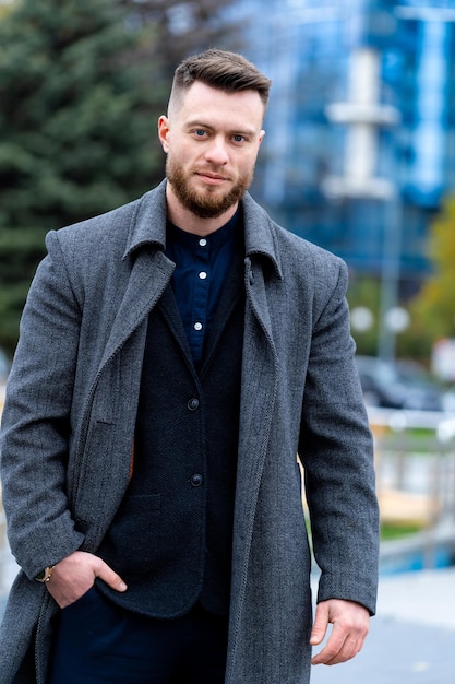 Stilvoller Mann, der die Hand am Mantel hält und in der Nähe von hohen Gebäuden spazieren geht Man entspannender Spaziergang auf der modernen Straße Lifestyle-Konzept