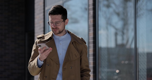 Stilvoller Mann, der Sprachnachrichten erstellt oder die App für virtuelle Assistenten auf dem Mobiltelefon verwendet Geschäftsmann mit Brille, der Sprachnachrichten über die Freisprecheinrichtung aufzeichnet, während er in der Nähe eines modernen Bürogebäudes geht