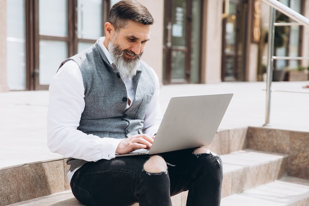 Stilvoller Mann des älteren Mannes mit grauem Bart verbringt Zeit draußen im Laptop.