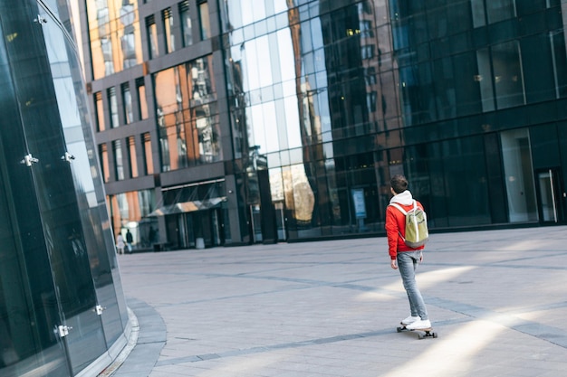 Stilvoller Mann in roter Jacke mit grünem Rucksack, der zwischen den modernen Häusern Spaß mit dem Skateboard hat