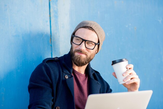 Stilvoller Mann, lässig gekleidet in Pullover und Hut, der mit Laptop arbeitet und mit Kaffee in der Nähe des blauen Wandhintergrunds sitzt