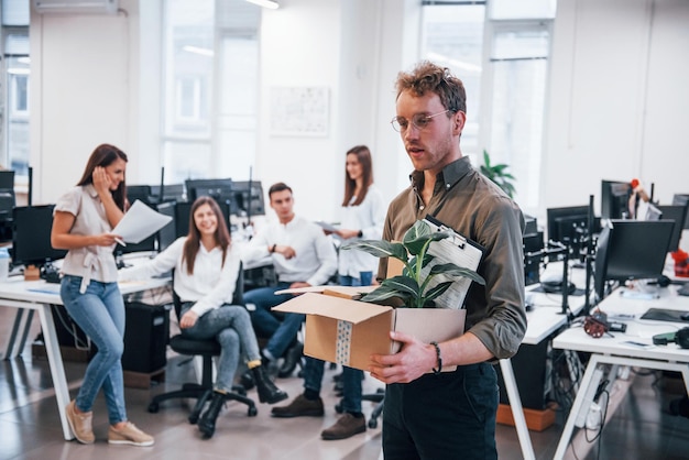 Stilvoller Mann mit Brille, der eine Kiste mit Pflanze vor seinen Kollegen im Büro hält.