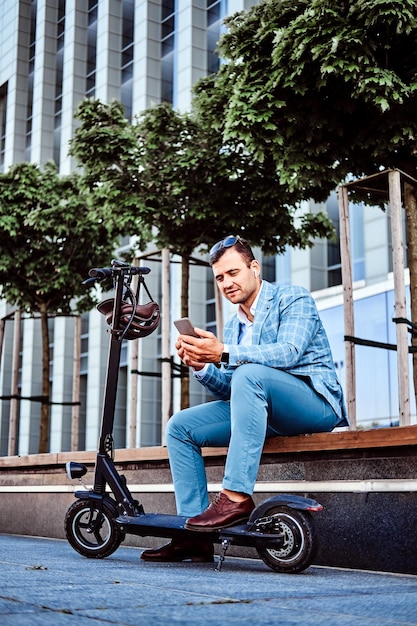 Stilvoller moderner Mann mit Elektroroller sitzt auf der Bank und plaudert auf seinem Handy.