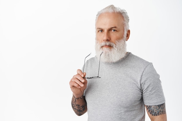 Foto stilvoller nachdenklicher alter mann, der die lippen mit brillenbügel berührt, nach vorne ernst schaut, nachdenkt, über eine entscheidung nachdenkt, in einem lässigen grauen t-shirt gegen eine weiße wand steht