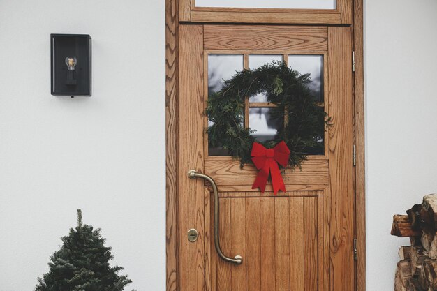 Stilvoller rustikaler Weihnachtskranz mit roter Schleife an Holztüren und Tannenbaum mit roter Schleife am Hauseingang. Winterliche Feiertagsdekoration der Fassade eines modernen Bauernhauses