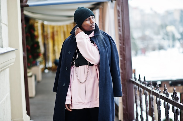 Stilvoller städtischer Artafroamerikanermann im schwarzen Mantel und im Hut, rosa Hoodie warf am Wintertag auf.