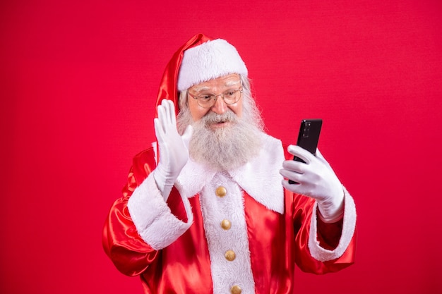 Stilvoller trendiger Großvater im Alter von reifem Weihnachtsmann-Traditions-Winter-Kostüm-Kopfbedeckungs-Brille weißer Bart nehmen Weihnachts-Selfie-Bild-Frontkamera in rotem Hintergrund.