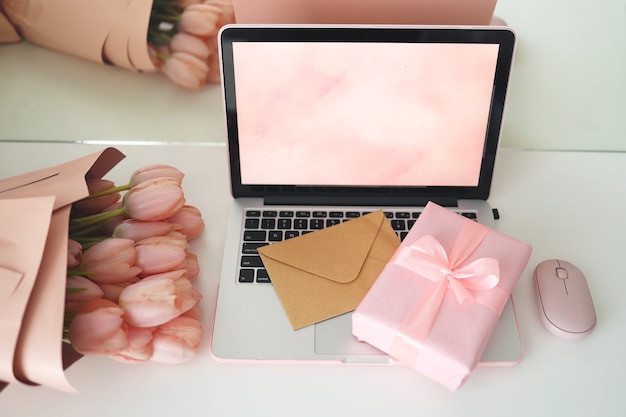 Stilvoller weiblicher Schreibtischarbeitsplatz mit rosa Tulpen, Laptop-Computer und Umschlag. Notebook-Bildschirm in Draufsicht. Rosa Geschenkbox und rosa Computermaus.