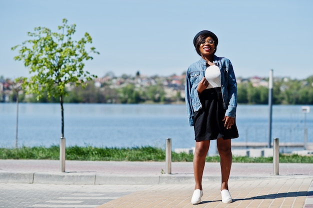 Stilvolles Afroamerikanermodell im Glashut, in der Jeansjacke und im schwarzen Rock warf im Freien auf.