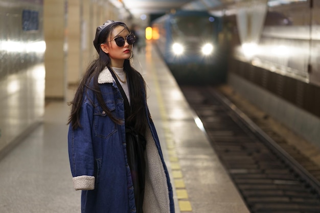 Stilvolles asiatisches Mädchen in Brille und Street-Fashion-Outfit am U-Bahnsteig mit ankommendem Zug