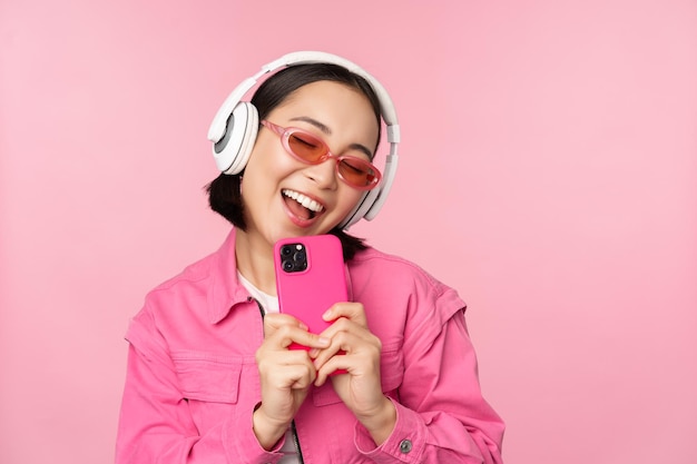 Stilvolles asiatisches Mädchen mit Kopfhörern, das Musik hört und Fotos auf dem Handy mit dem Smartphone macht, das über rosa Hintergrund steht