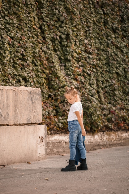 Stilvolles blondes kleines Mädchen in Jeans und weißem T-Shirt geht die Straße entlang. Mädchen 7 Jahre alt kleines Modell, schönes Kind