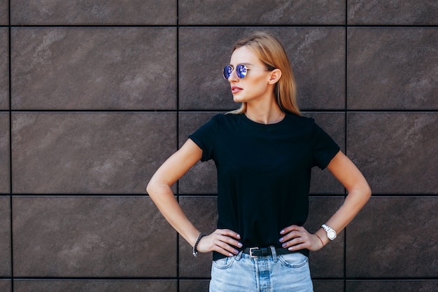 Stilvolles blondes Mädchen, das schwarzes T-Shirt und Brille trägt, die gegen Straße aufwirft