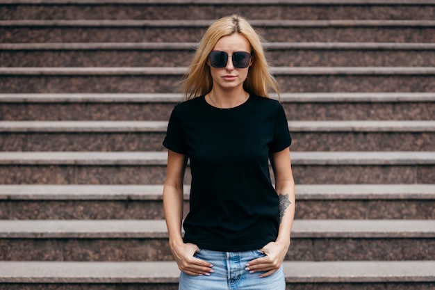Stilvolles blondes Mädchen, das schwarzes T-Shirt und Gläser trägt, die gegen Straße aufwerfen