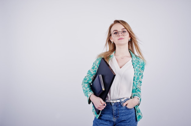 Stilvolles blondes Mädchen in der Jacke und in den Jeans an den Gläsern mit Notizbuchtagebuch und Laptop an den Händen gegen