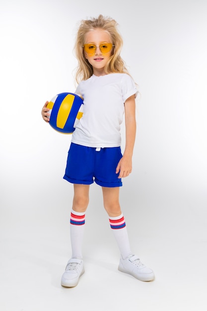 Stilvolles blondes Mädchen in Sportshorts, einem T-Shirt und Turnschuhen mit Brille hält einen Basketballball auf einem Weiß