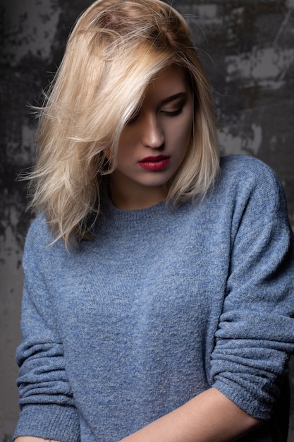 Stilvolles blondes Model mit üppigem Haar, das einen blauen Pullover trägt und im Studio mit Schatten posiert