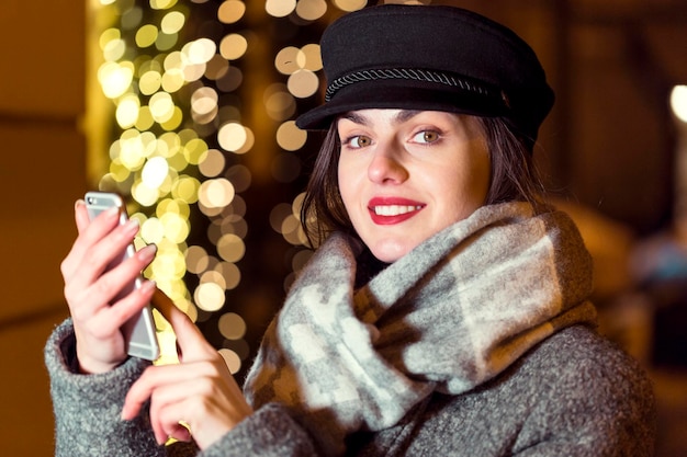 Stilvolles brünettes lächelndes Mädchen mit Smartphone vor der Girlandenwand in der Weihnachtszeit am Abend in der Stadt