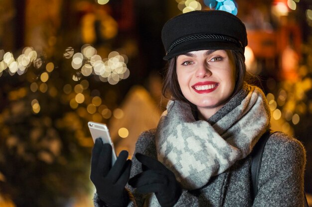 Stilvolles brünettes lächelndes Mädchen mit Smartphone vor der Girlandenwand in der Weihnachtszeit am Abend in der Stadt