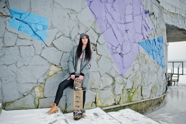 Stilvolles brünettes Mädchen im lässigen Streetstyle mit grauer Kappe und Skateboard am Wintertag gegen farbige Wand