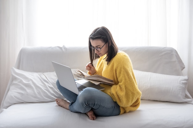 Stilvolles brünettes Mädchen in einem gelben Pullover sitzt zu Hause auf der Couch mit einem Buch und einem Laptop. Das Konzept des Studentenlebens und der Selbstentwicklung.