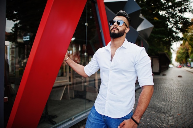 Stilvolles großes arabisches Mannmodell in weißer Hemdjeans und Sonnenbrille posierte auf der Straße der Stadt Bart attraktiver arabischer Kerl