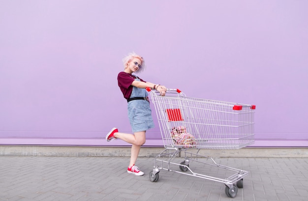 Stilvolles helles Mädchen mit gefärbten Haaren wirft mit einem Einkaufswagen auf einem lila Hintergrund auf