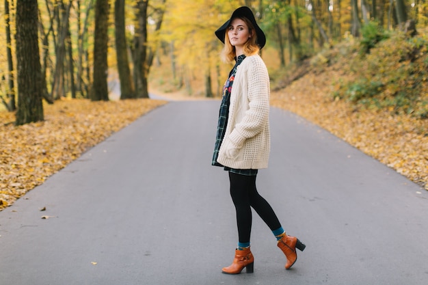 Stilvolles Hippiemädchen in einer gestrickten Strickjacke und in einem Hut geht in den Herbstpark.