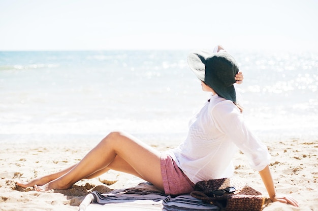 Stilvolles Hipster-Mädchen mit Hut, das am Strand sitzt und sich in der Nähe von Meereswellen bräunt Sommerurlaub Fröhliche Boho-Frau, die sich entspannt und einen sonnigen warmen Tag am Meer genießt Platz für Text