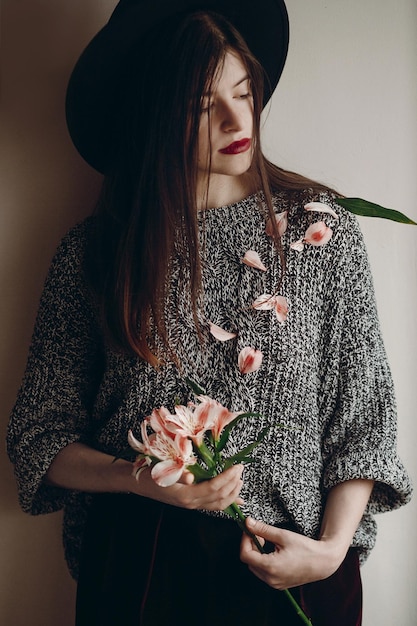 Stilvolles Hipster-Mädchen mit Hut, das rosa Blumen und Blütenblätter auf dem Pullover hält, wachsende Boho-Frau mit schöner Alstroemeria in den Händen, kreatives, sinnliches weibliches Porträt-Öko-Konzept, speichern und schützen