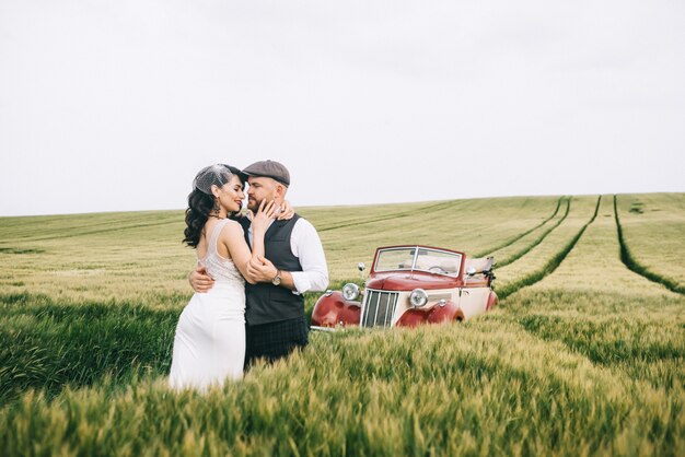 Stilvolles Hochzeitspaar in einem grünen Feld nahe Retroauto.