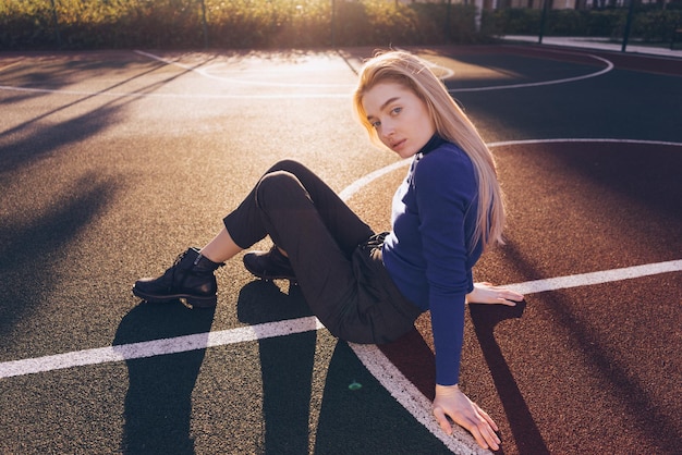 Stilvolles junges blondes Model-Mädchen im blauen Pullover sitzt auf dem Spielplatz und posiert